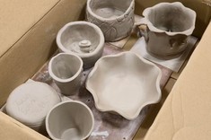 W kartonie umieszczone ulepione kubki i miski z gliny w stanie surowym w szarym kolorze.