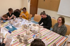 W niewielkim pomieszczeniu wokół stołu zgromadzonych jest kilka osób podczas wykonywania przedmiotów z gliny. 