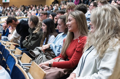 Zbliżenie na grupę studentów słuchających wykładu. W tle sala wykładowa wypełniona w całości słuchaczami wykładu.