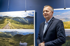 Na pierwszym planie stoi mężczyzna w ciemnej marynarce i zwraca się do zebranych osób. W tle na granatowej ścianie zawieszone plansze z dużymi kolorowymi fotografiami pejzaży górskich.