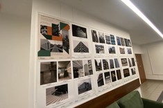 W pomieszczeniu na jasnej ścianie około dwudziestu zdjęć w stonowanych barwach rozplanowanych na prostokątnych planszach. W górnym lewym rogu kolorowy plakat wystawy. Ściana widoczna pod kątem.