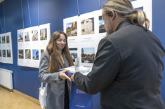 Mężczyzna ustawiony plecami do zdjęcia wręcza nagrodę młodej kobiecie z długimi włosami.  W tle plansze ze zdjęciami.