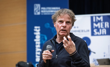 Opis: Mężczyzna, prelegent ubrany na czarno, podczas prowadzenia wykładu. W tle biało-niebieskie banery informacyjne.