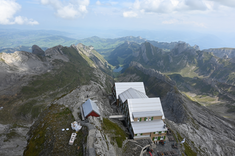 Zdjęcie wykonane z lotu ptaka. Zdjęcie budynku na szczycie wzniesienia. W tle surowy krajobraz wysokich gór.