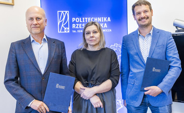Na tle białej ściany z niebieskim pasem z napisem politechnika rzeszowska stoją obok siebie trzy osoby dwóch mężczyzn i kobieta po środku. Wszyscy elegancko ubrani w różnych odcieniach niebieskiego. Uśmiechają się. Mężczyźni trzymają w rękach okładki z dokumentami.