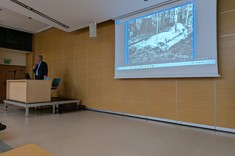 Na zdjęciu na tle ściany z jasnym drewnopodobnym wykończeniem stoi po lewej stronie na niewielkim podwyższeniu za jasnym biurkiem prelegent, mężczyzna w ciemnej marynarce. Po prawej stronie widoczny duży ekran z wyświetloną prezentacją.