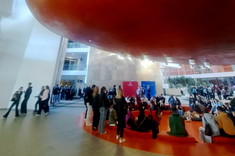 W jasno oświetlonym dużym wnętrzu holu po środku na wyznaczonym na posadzce czerwonym kole zgromadzonych dużo młodych osób. Nad kołem zawieszona wysoko duża czerwona bryła o obłym kształcie.