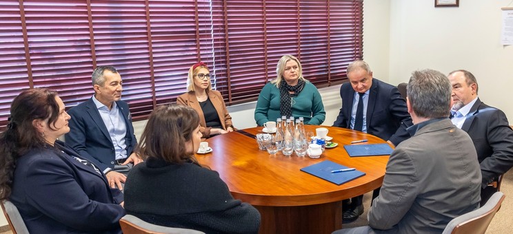 Na zdjęciu po środku pomieszczenia jasno brązowy okrągły drewniany stół. Wokół stołu siedzi osiem osób podczas spokojnej rozmowy, cztery kobiety i czterech mężczyzn. Wszyscy elegancko ubrani.