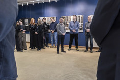 We wnętrzu z ciemnoniebieskimi ścianami, na tle plansz wywieszonych na ścianie, stoi grupa kilkunastu osób.