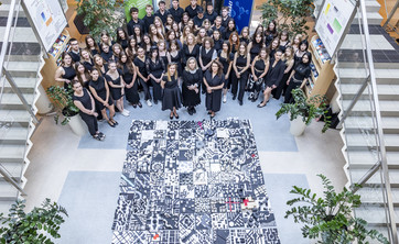 Zdjęcie wykonane z góry. W centrum ujęcia na podłodze ułożony kwadrat około 3 x 3 metry z biało-czarnych kwadratowych makiet. Wokół zgromadzona grupa kilkudziesięciu osób. wszyscy ubrani na czarno.