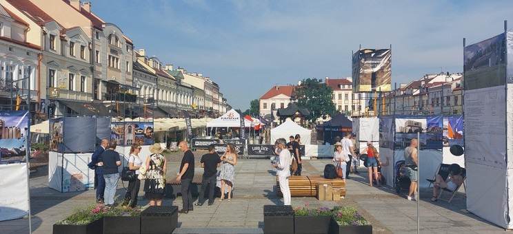 Zdjęcie przedstawia większy fragment Rynku w Rzeszowie. Po środku donice z kwiatami i drewniane ławki. Po bokach ustawione plansze. Na środku grupa ludzi rozmawiających ze sobą.