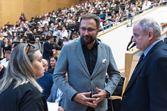 Trzy osoby, kobieta i dwóch mężczyzn, stoją zwrócone do siebie podczas rozmowy. W tle uczestnicy wykładu.