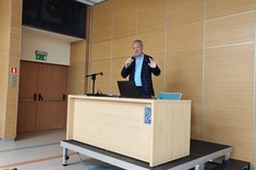 Na zdjęciu na tle ściany z jasnym drewnopodobnym wykończeniem stoi na niewielkim podwyższeniu za jasnym biurkiem prelegent, mężczyzna z siwymi włosami w ciemnej marynarce i niebieskiej koszuli.