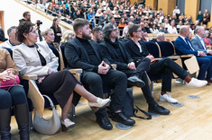 Duża grupa osób siedzących w rzędach podczas wykładu. Na pierwszym planie, pośrodku, trzech mężczyzn ubranych na czarno. 