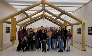 W jasnym wnętrzu pośrodku ustawiony szkielet konstrukcji z papierowych tub przypominający dom z wysokim dachem. Pod konstrukcją grupa około dwudziestu osób pozujących do zdjęcia.