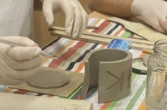 Zbliżenie na ręce w trakcie pracy nad przedmiotem z gliny. Na zdjęciu widoczny walec z gliny z wyrzeźbionym napisem i kolista podstawa.