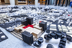 Zbliżenie na kilka biało-czarnych makiet. Każda z makiet jest wykonana na podstawie kwadratowej. Pośrodku jedyna makieta z akcentem koloru czerwonego.