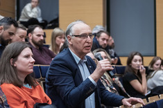 Zbliżenie na słuchaczy wykładu siedzących w rzędach. Po środku uczestnik wykładu, mężczyzna w ciemnej marynarce podczas zadawania pytania prelegentowi.