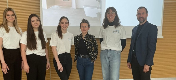 Na tle ekranu z wyświetloną prezentacją grupa sześciu osób. Po lewej mężczyzna w ciemnym ubraniu. Pośrodku grupy kobiet w białych podkoszulkach prelegentka w ciemnym ubraniu. Wszyscy się uśmiechają.