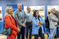 Ujęcie pomieszczenia, w którym stoi grupa kilku osób. Wszyscy zwróceni w jednym kierunku, uśmiechają się. W tle na granatowej ścianie zawieszone plansze z dużymi kolorowymi fotografiami.