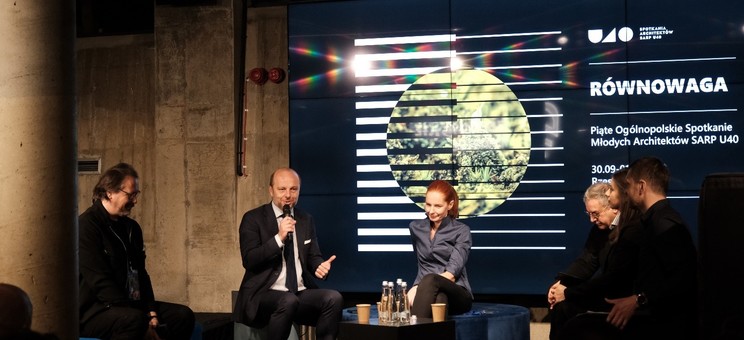 Pomieszczenie z betonowymi ścianami, kameralnie oświetlone. Pośrodku scena, na której jest kilka siedzących na sześcianach osób. W tle zdjęcie z geometryczną grafiką i napisem U40- Równowaga.