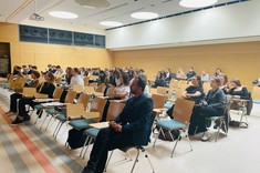 W jasnym wnętrzu sali wykładowej ze ścianami z drewnopodobną wykładziną i z białym sufitem zgromadzona kilkudziesięcioosobowa grupa słuchaczy. Wszyscy zajmują miejsca siedzące.