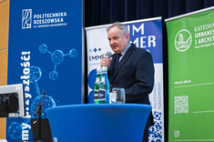 Mężczyzna w ciemnym garniturze stoi za niebieskim pulpitem, przemawia do zebranych.  W tle biało-niebieskie i zielono-białe banery informacyjne jednostek politechniki.  
