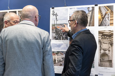 Mężczyzna w ciemnym garniturze wskazuje dłonią na fragment wywieszonego zdjęcia, a dwóch mężczyzn obok przygląda się fotografii.