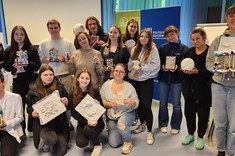 W pomieszczeniu zgromadzonych jest razem kilkanaście osób. Jedni stoją, drudzy przysiadają, wszyscy pozują razem do zdjęcia i trzymają w rękach różne prace plastyczne. Wszyscy się uśmiechają.