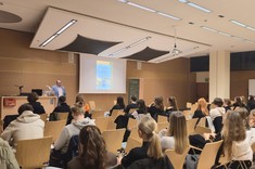 Pomieszczenie z białym sufitem i drewnopodobnym wykończeniem ścian. Po środku przy podwyższeniu stoi mężczyzna podczas prowadzenia wykładu. W tle ekran z wyświetlonym slajdem.  