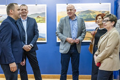 W pomieszczeniu stoi obok siebie pięć osób, trzech mężczyzn po lewej stronie i dwie kobiety po prawej. Rozmawiają ze sobą i uśmiechają się. W tle na granatowej ścianie zawieszone plansze z dużymi kolorowymi fotografiami.