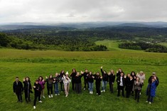 Zdjęcie wykonane z wysokości kilkudziesięciu metrów. Grupa około dwudziestu osób ubranych na sportowo pozuje do zdjęcia. Wszyscy spoglądają do góry i machają do obiektywu. W tle zielona polana i panorama gór.