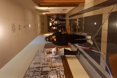 Zdjęcie wewnątrz budynku, na ścianach widoczne planszhttps://zuia.prz.edu.pl/filese z pracami studentów, na podłodze znajduje się wyznaczona trasa, po której chodzi kobieta w opasce na oczy oraz laską dla niewidomych. W tle znajduje się grupa ludzi. 