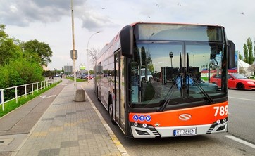 Na kolorowym zdjęciu po środku autobus stojący na przystanku. Po lewej stronie widoczny chodnik i ścieżka rowerowa, w tle zieleń.