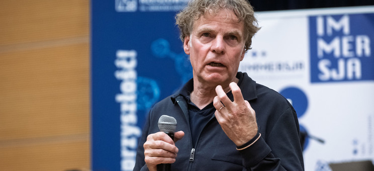 Opis: Mężczyzna, prelegent ubrany na czarno, podczas prowadzenia wykładu. W tle biało-niebieskie banery informacyjne.