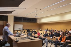 W pomieszczeniu z przyciemnionym światłem, po lewej stronie za stołem stoi mężczyzna prowadzący wykład. Na przeciwko siedzi grupa ponad dwudziestu osób, słuchaczy wykładu.          