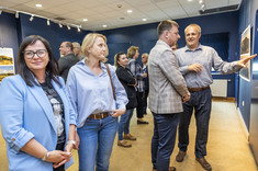 W pomieszczeniu na pierwszym planie stoją dwie kobiety. Obie ubrane na niebiesko. Jedna uśmiecha się do zdjęcia.  W tle dwóch mężczyzn stoi przed prezentowanymi planszami i wskazują na zdjęcia.