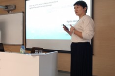 Na zdjęciu ekran do wyświetlania prezentacji, przed nim biały stół. Po prawej stronie zdjęcia kobieta prezentująca slajdy z ciemnymi krótkimi włosami. Ubrana jest w długą czarną spódnicę i białą bluzkę.