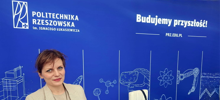 Na pierwszym planie kobieta w jasnej marynarce uśmiecha się do zdjęcia. W rękach trzyma folder reklamowy politechniki.  W tle ciemno ciemno niebieska ściana z białymi rysunkami i wyraźnymi napisami: Politechnika Rzeszowska i Budujemy przyszłość