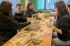 W pomieszczeniu przy długim stole siedzą cztery osoby, trzy kobiety i jeden mężczyzna. Wszyscy ubrani na ciemno. Na stole rozłożone różne materiały, zgromadzeni wykonują z nich prace plastyczne.