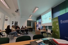 W lekko przyciemnionym pomieszczeniu widocznych jest po lewej stronie kilka osób słuchających wykładu. Po prawej stronie wysoko zawieszony jasny ekran, przed nim siedzi przy stole prelegentka.