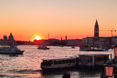 Zdjęcie w ciepłych barwach pokazujące zachód słońca nad miastem. Zdjęcie wykonane z oddali, przed miastem widoczna zatoka z kilkoma łodziami.