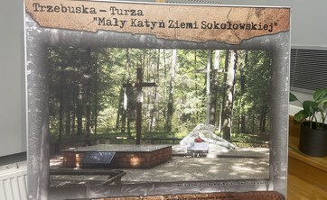 Na zdjęciu widoczny jest plakat przedstawiający miejsce pamięci w lesie turzańskim, nazwane „Mały Katyń Ziemi Sokołowskiej”. Centralnym punktem fotografii jest pomnik z krzyżem, otoczony drzewami. Na pomniku znajduje się tablica pamiątkowa, a przed nim widoczne są kwiaty i znicze.