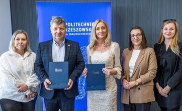 Od lewej prof. A. Sikora, rektor, prof., P. Koszelnik, dr. B. Szafrańska, p. P. Multan, arch. D. Bober. Na zdjęciu na tle ciemnej ściany z wyróżniającym się niebieskim banerem z białym napisem Politechnika Rzeszowska stoi pięć elegancko ubranych osób. Dwie osoby pośrodku trzymają okładki z umowami.