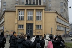 Zdjęcie na ulicy, przed monumentalnym historycznym budynkiem stoi w luźnym układzie grupa około dwudziestu osób.
