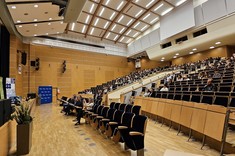 Zdjęcie przedstawia przestronną i nowoczesną salę audytoryjną podczas trwającego wydarzenia. Po lewej stronie, na scenie , stoi mężczyzna przemawiający do mikrofonu. Widownia jest częściowo zapełniona – uczestnicy siedzą w eleganckich, drewnianych fotelach z granatową tapicerką, a ich uwaga skupiona jest na prelegencie. Sala wyróżnia się nowoczesnym designem z drewnianymi panelami na ścianach i suficie, oraz starannie rozmieszczonym oświetleniem.