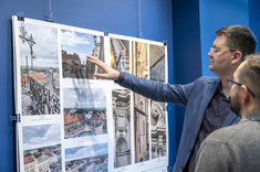 Mężczyzna w ciemnym garniturze wskazuje dłonią na fragment wywieszonego zdjęcia, a mężczyzna obok przygląda się fotografii.