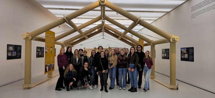 W jasnym wnętrzu pośrodku ustawiony szkielet konstrukcji z papierowych tub przypominający dom z wysokim dachem. Pod konstrukcją grupa około dwudziestu osób pozujących do zdjęcia.