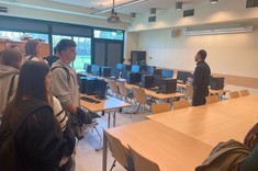 Na zdjęciu widać grupę uczniów w sali komputerowej. Stoją i słuchają mężczyzny, który prowadzi zajęcia. W tle są komputery na stołach, duże okna wpuszczają światło dzienne, a ściany są jasne.