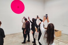 W jasnym wnętrzu grupa kilku osób stoi w jednej linii za sobą i każdy z nich robi zdjęcie dużemu czerwonemu kołu na białej ścianie.